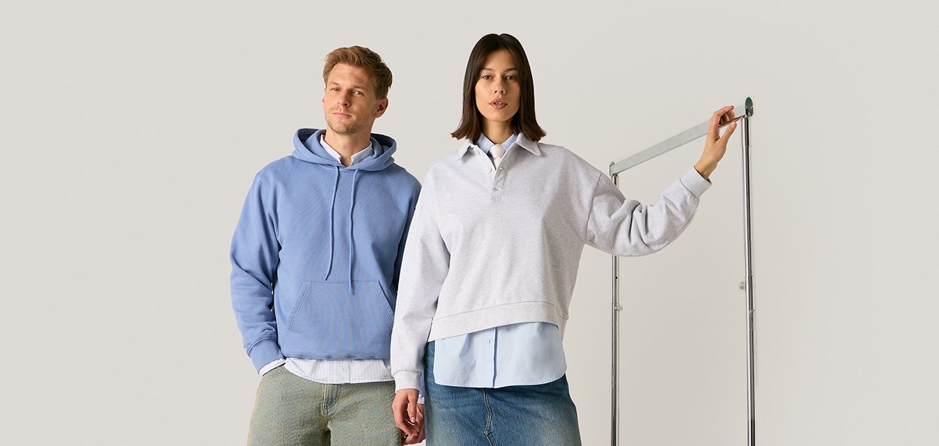 Mann in blauem Kapuzenpullover und hellen Hosen steht neben einer Frau im grauen Pullover und blauem Jeansrock mit Schlitz und hält eine Kleiderstangenstange.