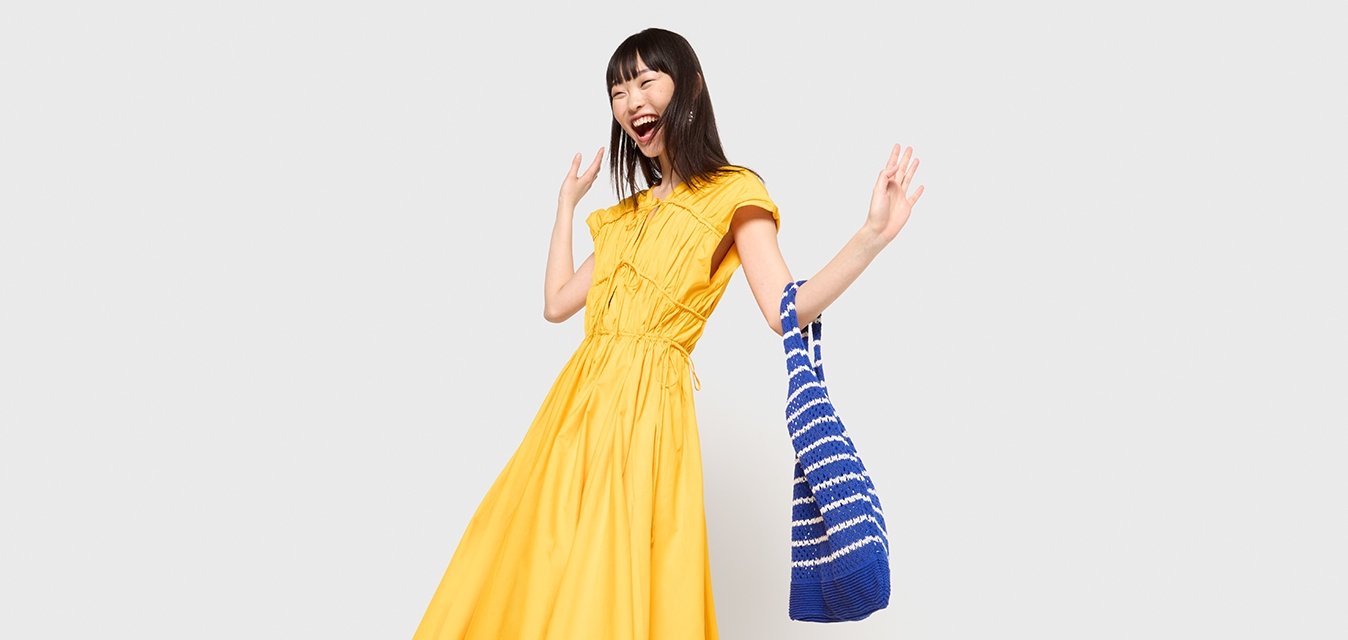 Gelbes, kurzärmliges, knöchellanges Kleid mit gerafften Details und Kordelzug in der Taille, kombiniert mit einer blau-weiß gestreiften Tasche.