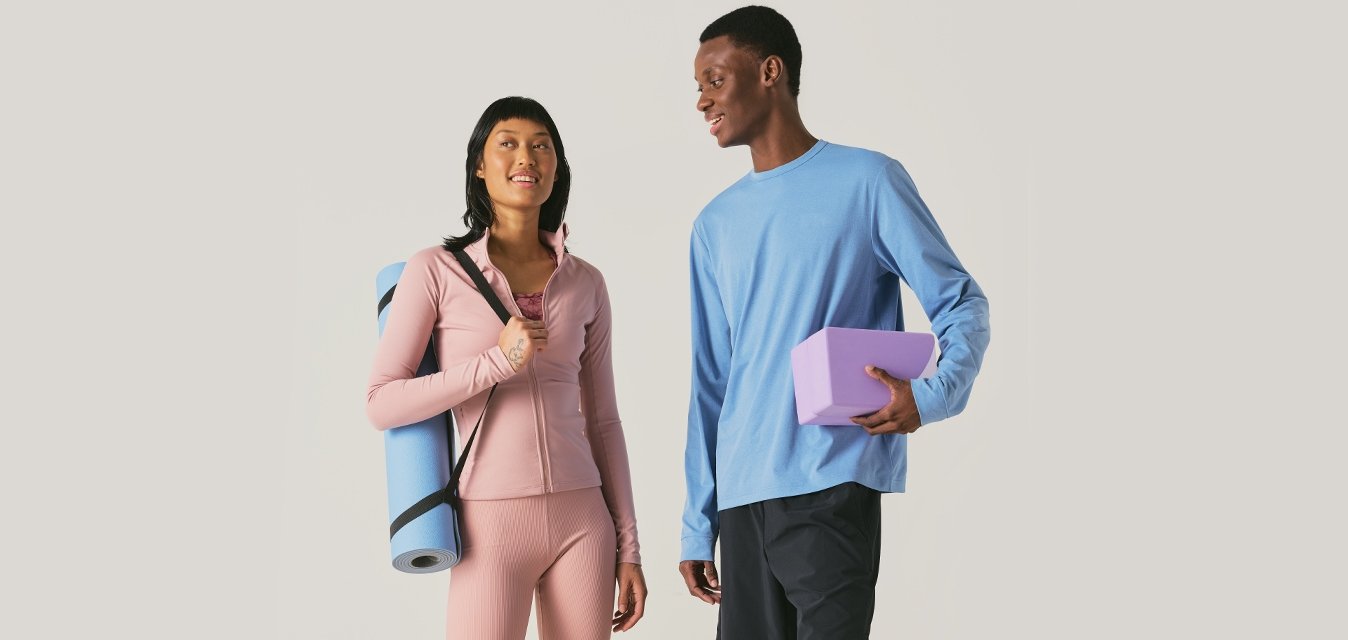Zwei Personen in Sportbekleidung, eine hält eine zusammengerollte Yogamatte, die andere hält einen Yogablock, beide lächeln und stehen zusammen.