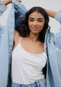 Frau mit dunklem, lockigem Haar, die ein weißes Tanktop und eine hellblaue Jeansjacke trägt und die Jacke von den Schultern hebt.
