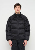 Mann trägt eine schwarze gesteppte Steppjacke mit hohem Kragen und schwarze Jogginghose, steht vor einem schlichten hellen Hintergrund.
