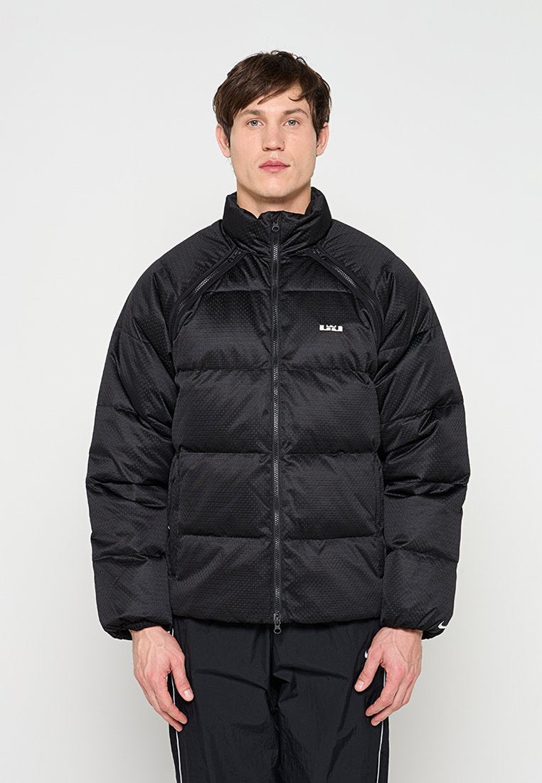 Mann trägt eine schwarze gesteppte Steppjacke mit hohem Kragen und schwarze Jogginghose, steht vor einem schlichten hellen Hintergrund.