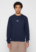 Junger Mann mit lockigem Haar trägt einen navyfarbenen Sweatshirt mit kleinem Logodruck auf der Vorderseite und beige Hosen, steht vor einem schlichten hellen Hintergrund.