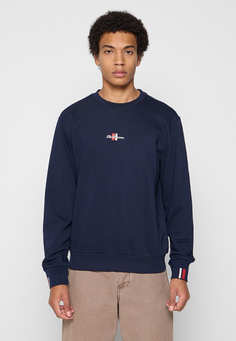 Junger Mann mit lockigem Haar trägt einen navyfarbenen Sweatshirt mit kleinem Logodruck auf der Vorderseite und beige Hosen, steht vor einem schlichten hellen Hintergrund.