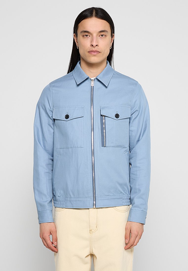 Helleblaue Zip-Jacke mit spitzem Kragen, zwei Fronttaschen und einer Reißverschlusstasche. Aus glattem Stoff gefertigt, verfügt sie über einen elastischen Bund.