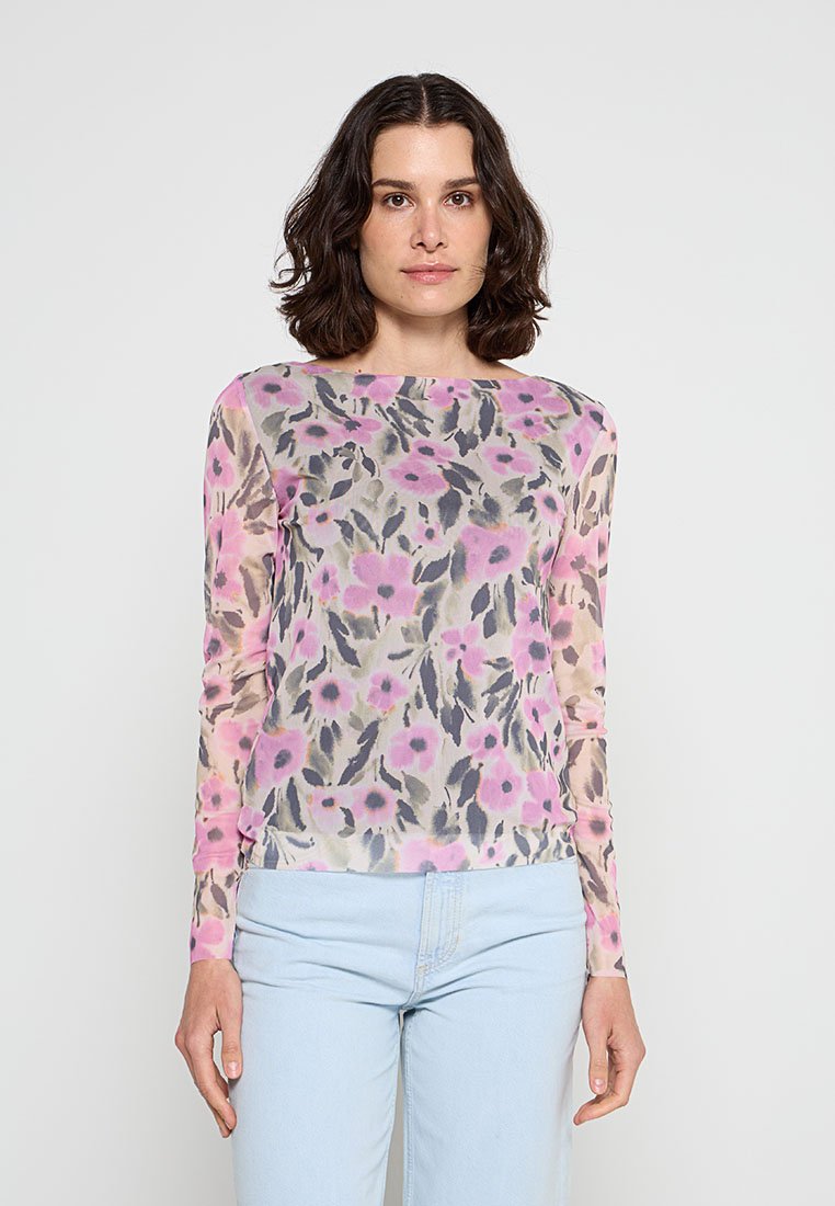 Frau mit dunkel schulterlangem Haar, die ein durchsichtiges langärmeliges Oberteil mit rosa-grauem Blumenmuster und hellblaue Jeans vor einfarbigem Hintergrund trägt.