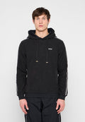 Junger Mann trägt schwarzen Hoodie mit Kordelzug vorne und schwarze Hose mit weißen Streifen an Ärmeln und Seiten, steht vor einem einfarbigen Hintergrund.