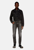 Mann mit geflochtenem Haar, der eine schwarze Denimjacke, ausgewaschene schwarze Jeans mit Abnutzungsdetails und schwarze Lacklederschuhe mit Metallkappen trägt.