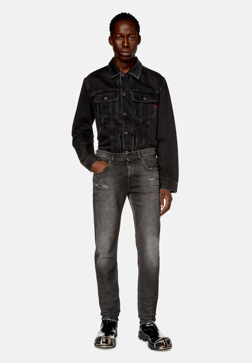 Mann mit geflochtenem Haar, der eine schwarze Denimjacke, ausgewaschene schwarze Jeans mit Abnutzungsdetails und schwarze Lacklederschuhe mit Metallkappen trägt.