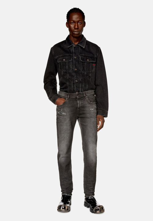Mann mit geflochtenem Haar, der eine schwarze Denimjacke, ausgewaschene schwarze Jeans mit Abnutzungsdetails und schwarze Lacklederschuhe mit Metallkappen trägt.