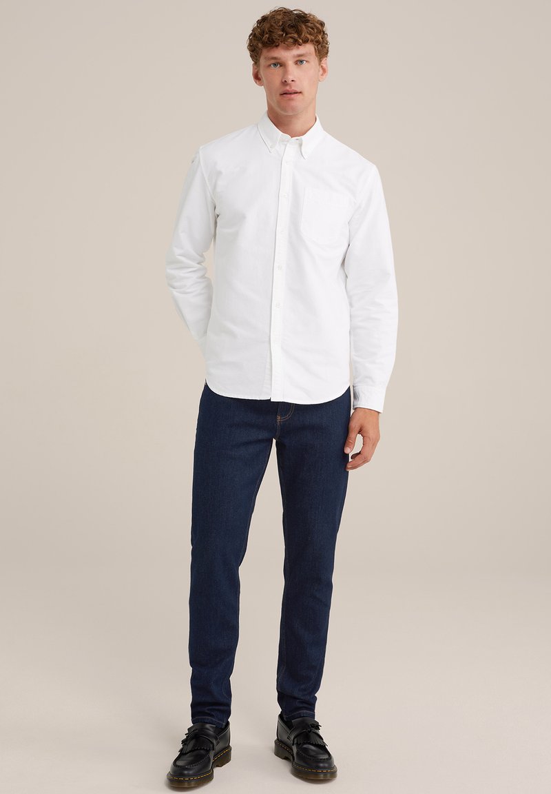 Weißes Baumwollhemd mit Knöpfen und Brusttasche, kombiniert mit dunkelblauen Jeans und schwarzen Schuhen mit Fransen. Glatte Textur insgesamt.