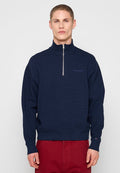 Mann trägt einen marineblauen Pullover mit halbem Reißverschluss und rote Hose, steht vor einem schlichten hellen Hintergrund, schaut nach vorne mit neutralem Gesichtsausdruck.