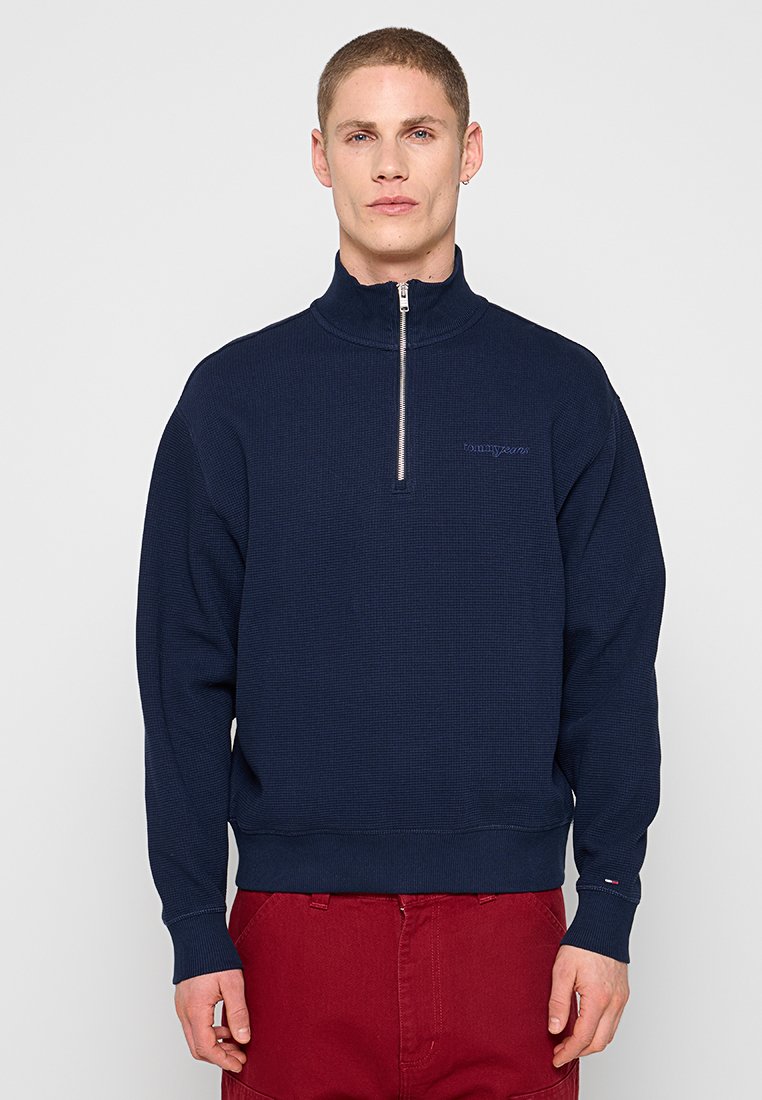 Mann trägt einen marineblauen Pullover mit halbem Reißverschluss und rote Hose, steht vor einem schlichten hellen Hintergrund, schaut nach vorne mit neutralem Gesichtsausdruck.