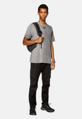 Graues, verblichenes T-Shirt mit lockerer Passform; schwarze Cargo-Hosen mit Taschen; schwarzer Rucksack; schwarze Sportschuhe mit strukturierten Sohlen; silberne Uhr.