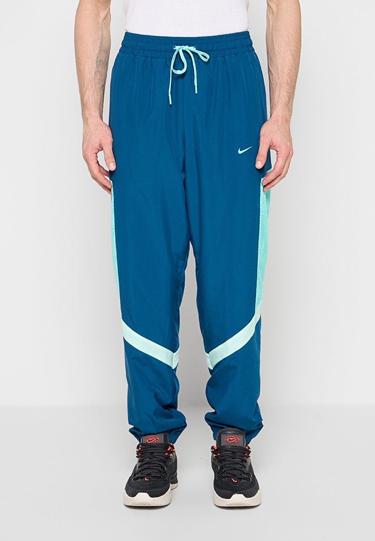 Mann trägt blaue Nike-Trainingshose mit hellblauen Streifen und schwarzen Turnschuhen, steht vor einem einfarbigen weißen Hintergrund.