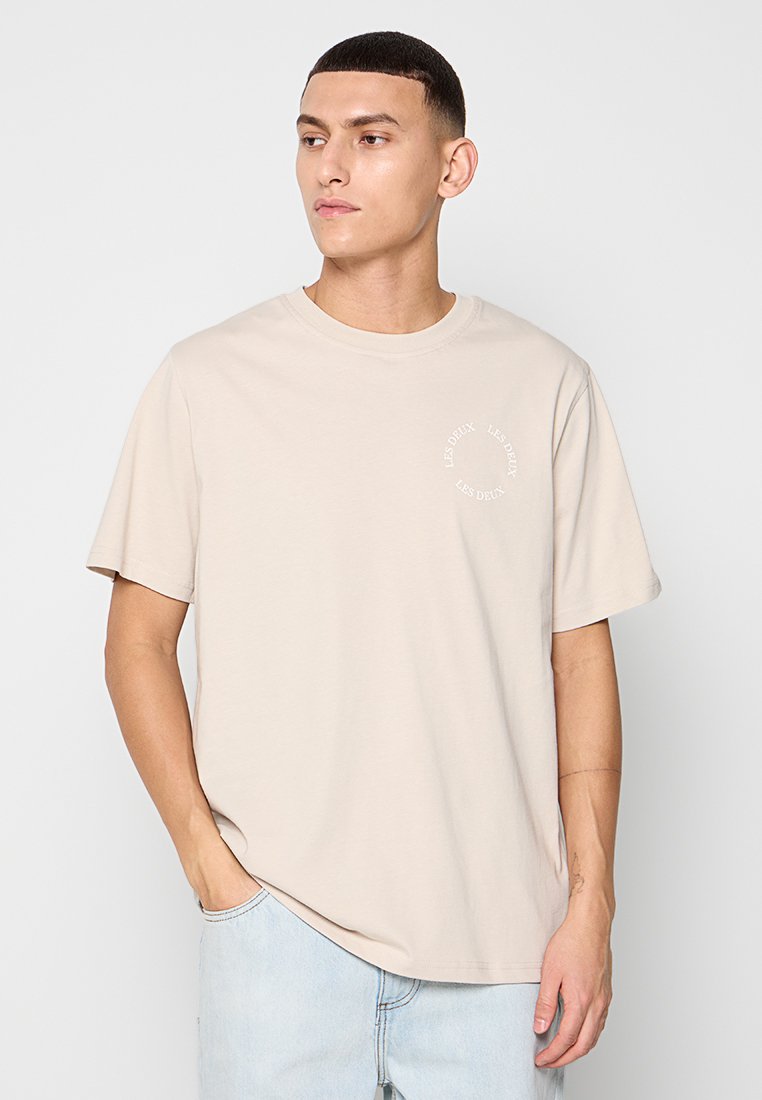 Beige Rundhals-T-Shirt aus Baumwolle, mit kurzen Ärmeln und einem bedruckten runden Logo auf der linken Brust. Glatte Textur, lässige Passform.