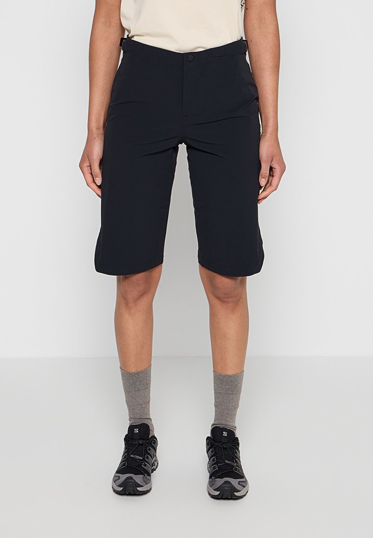 Person trägt schwarze knielange Shorts, graue Socken, schwarze und graue Sportschuhe, steht vor einem einfachen hellen Hintergrund.
