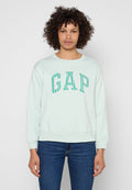 Person mit lockigem Haar, der ein hellmintgrünes Sweatshirt mit "GAP" in türkis und pfirsichfarben gestreiften Buchstaben trägt, kombiniert mit blauen Jeans.