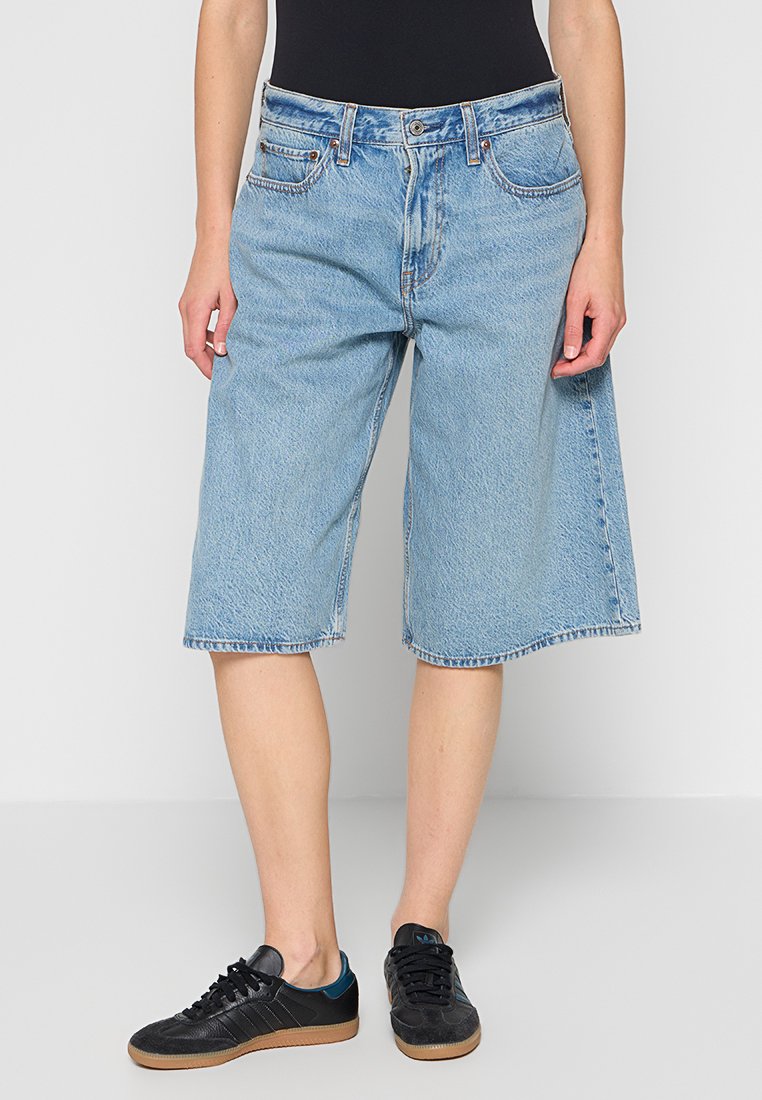 Hellblaue Denim-Shorts mit lockerem Schnitt, mittelhoher Taille und rohem Saum. Merkmale klassische Nähte und ein Design mit fünf Taschen. Getragen mit schwarzen Sneakern.