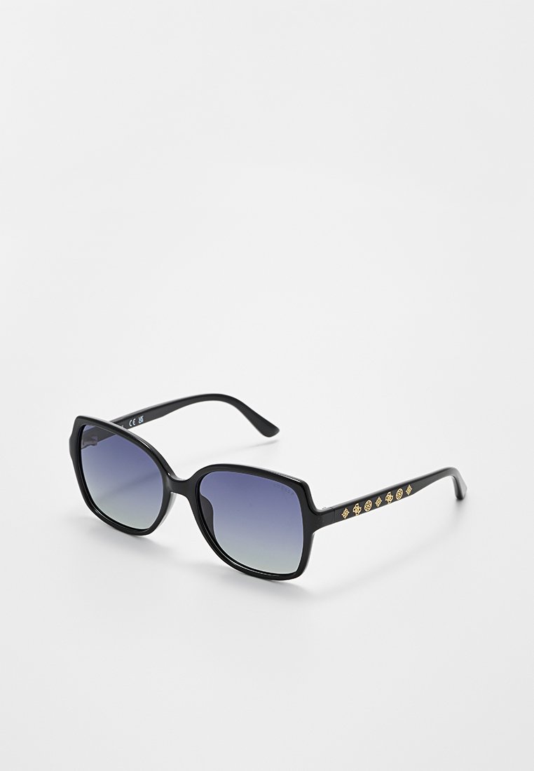 Schwarze übergroße quadratische Sonnenbrille mit Verlauf blau getönten Gläsern und goldenen dekorativen Symbolen an den Bügeln, auf weißem Untergrund gezeigt.