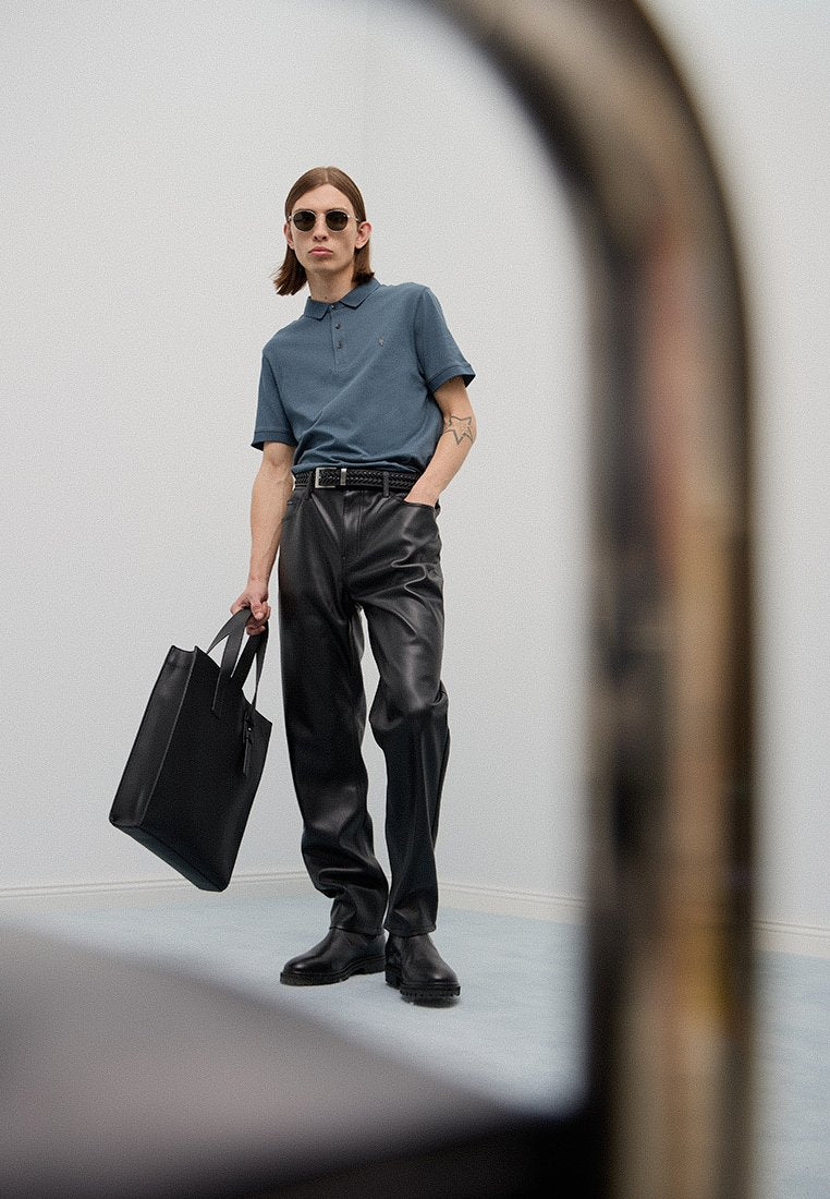 Dunkelblaues Poloshirt aus Stoff, schwarze Lederhose, schwarze Schuhe und eine schwarze Tragetasche. Schlank geschnittenes Design mit minimalistischem Stil und Sonnenbrille.