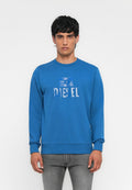Mann trägt einen blauen Diesel-Pullover und graue Jeans, steht vor einem schlichten weißen Hintergrund, blickt nach vorne mit neutralem Ausdruck.