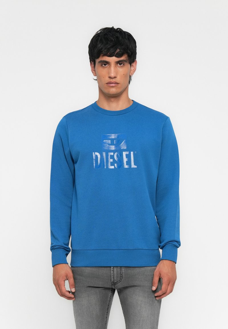 Mann trägt einen blauen Diesel-Pullover und graue Jeans, steht vor einem schlichten weißen Hintergrund, blickt nach vorne mit neutralem Ausdruck.