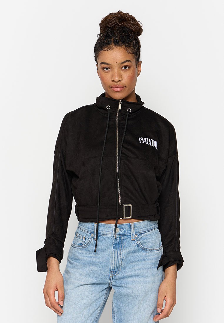 Junge Frau mit einem Haarknoten, getragen in einer schwarzen, croptop Jacke mit dem Schriftzug "PEGADO" und hellblauen, hochgeschnittenen Jeans, die vor einem weißen Hintergrund steht.