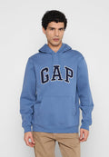 Mann mit lockigem schwarzem Haar, der einen blauen GAP-Hoodie und beige Hose trägt und vor einem schlichten hellgrauen Hintergrund steht.