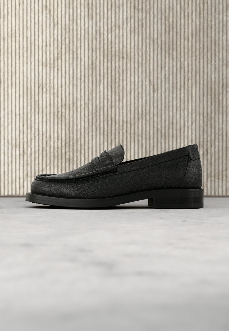 Schwarzer Leder-Herrenschuh mit abgerundetem Zehenbereich und niedrigem Absatz, auf einem glatten Boden vor einem strukturierten Hintergrund mit vertikalen Linien dargestellt.
