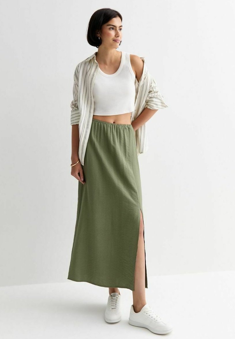 Lange olivgrüne Rock mit seitlichem Schlitz, kombiniert mit einem weißen Crop-Top und einem gestreiften Hemd, accessorized mit weißen Sneakers.
