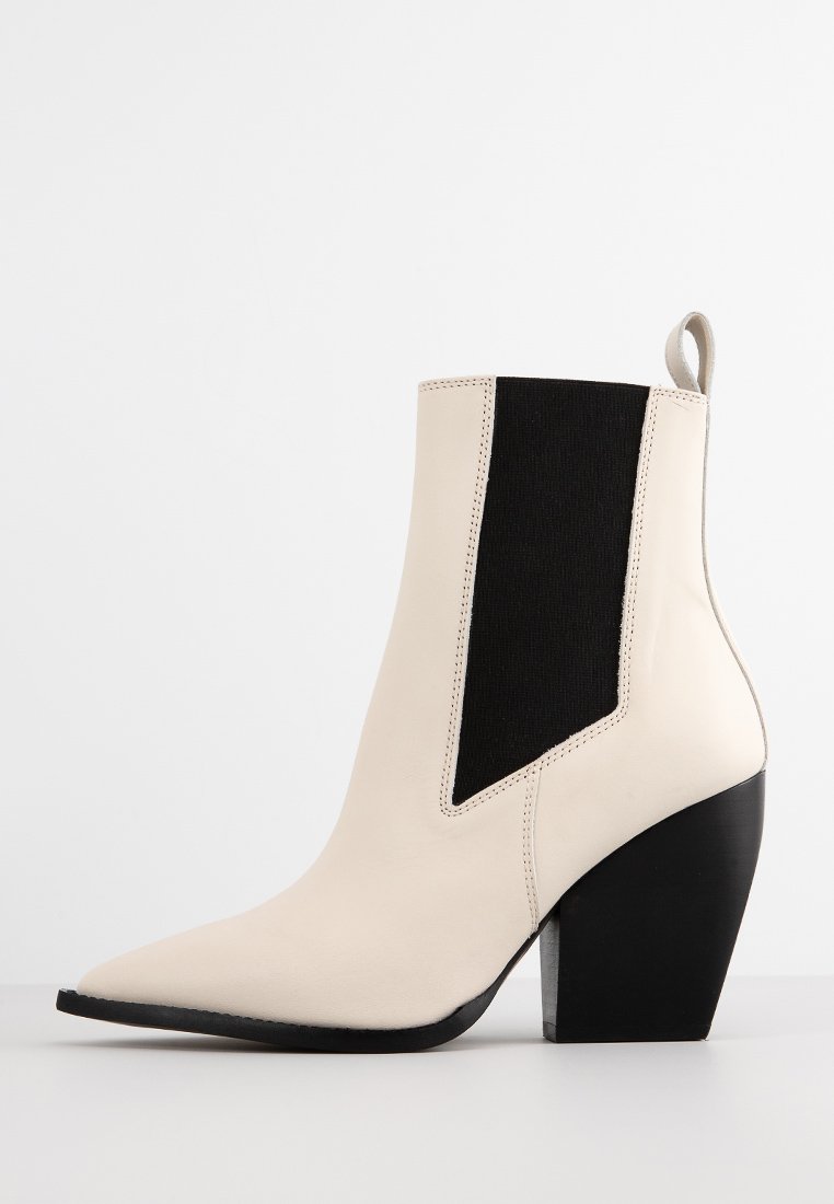 Cremfarbene Leder-Stiefelette mit schwarzem elastischem Seitenpanel und klobigem schwarzen Absatz auf weißem Hintergrund.