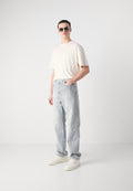 Hellbeiges T-Shirt mit kurzen Ärmeln, hoch taillierte Jeans in Blau und Weiß gestreift, und weiße Turnschuhe mit strukturiertem Schnürsenkel-Detail. Schwarze Sonnenbrille.