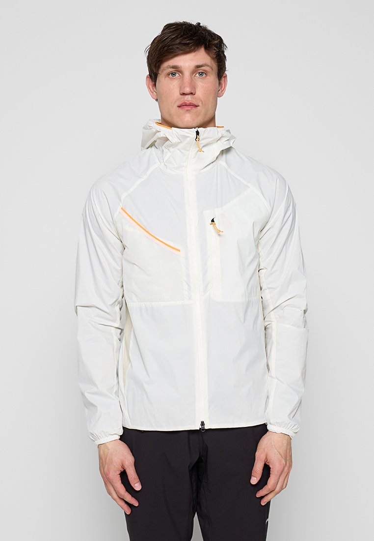 Mann trägt eine weiße, leichte Kapuzenjacke mit orangenen Reißverschlussakzenten und schwarze Hose, steht vor einem neutralen Hintergrund.