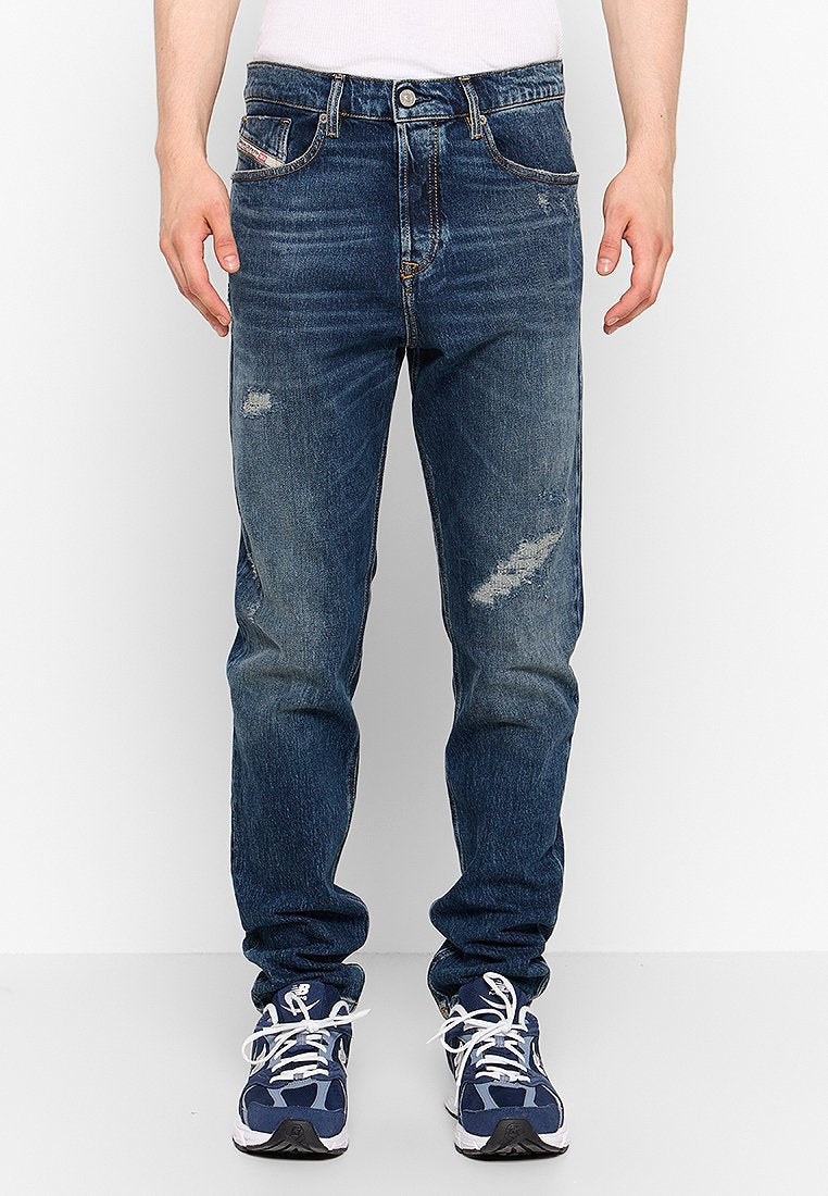 Dunkelblaue Jeans aus Denim mit Knopfverschlus, verblassten Stellen und Abnutzungen, fünf Taschen und geradem Bein. Getragen mit navyfarbenen Sneakers.