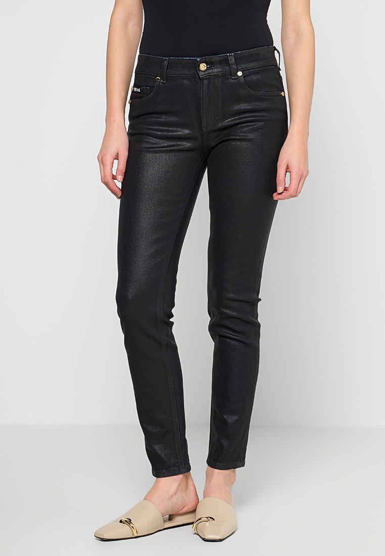Schwarze, glänzende Slim-Fit-Jeans mit goldenem Zubehör, fünf Taschen und einer mittelhohen Taille, kombiniert mit beigen Slippers mit einem goldenen Akzent.