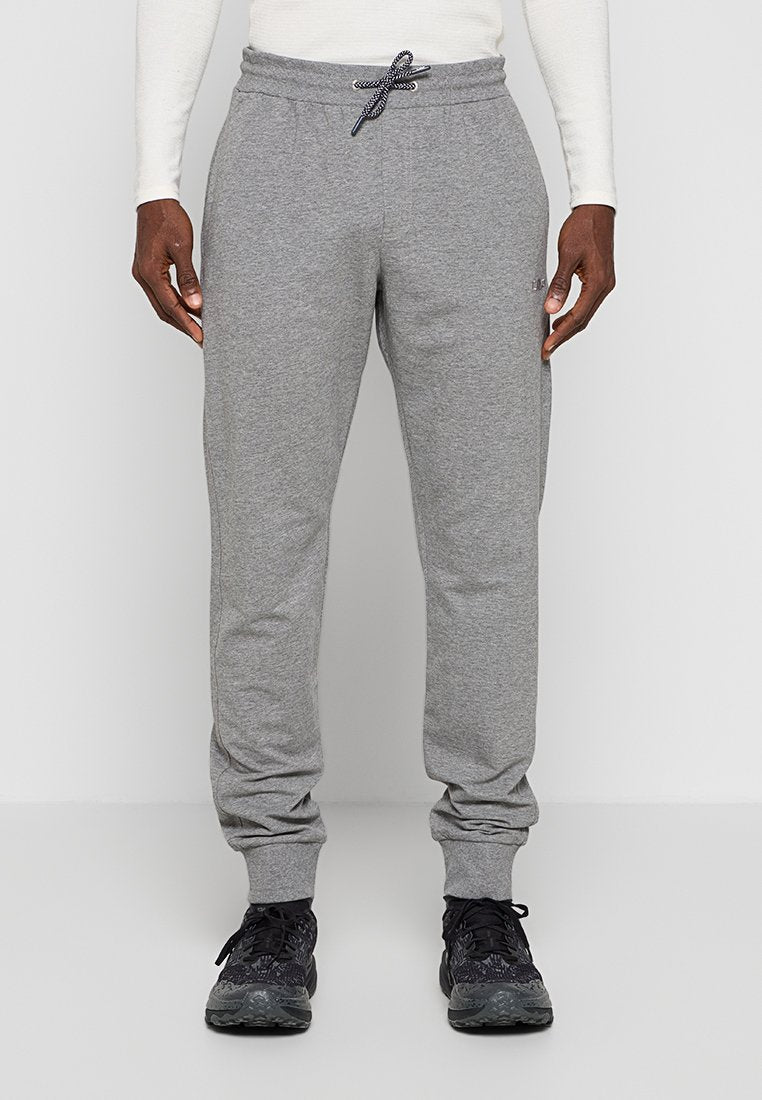 Mann trägt graue Jogginghose mit Kordelzug und Bündchen an den Knöcheln und schwarze Sneakers, steht vor einem hellen, einfachen Hintergrund.