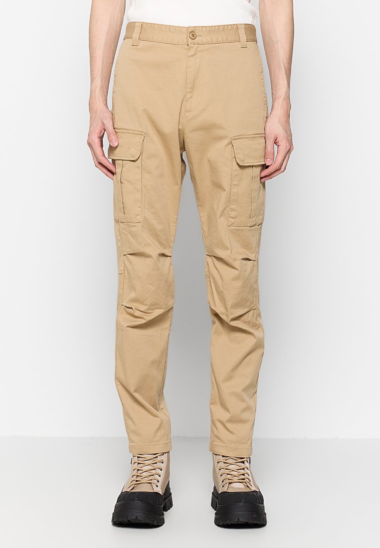 Beige Cargo-Hosen aus Baumwolle mit zwei frontalen Taschen und einer taillierten Passform. Getragen mit beigen Schnürboots und einem schlichten weißen Oberteil.