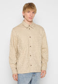 Beige Hemd mit Knopfleiste und subtiler gemusterter Textur, ausgestattet mit zwei Brusttaschen und braunen Knöpfen, kombiniert mit hellblauen Jeans.