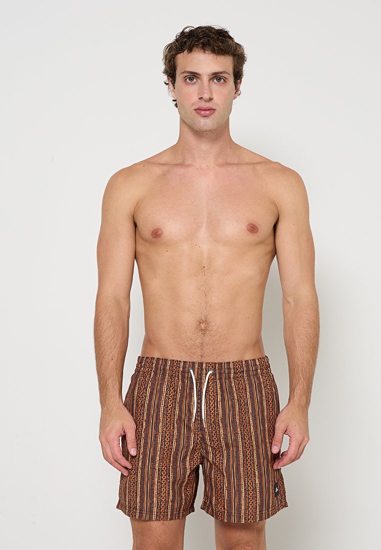 Junger Mann, der vornstehend braune gemusterte Badeshorts mit weißem Kordelzug trägt, vor einfachem hellem Hintergrund.