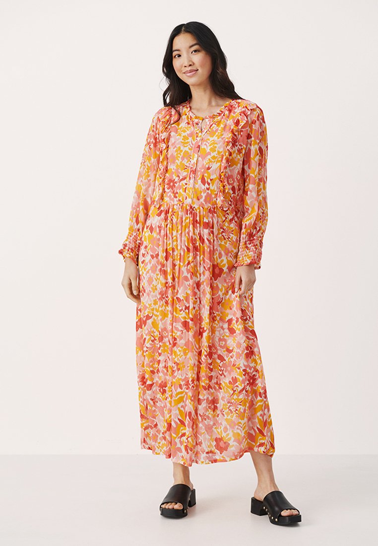 Florales Maxikleid in Pink-, Gelb- und Orangetönen. Lange Ärmel, geraffte Manschetten und eine taillierte Taille sorgen für eine fließende Silhouette. Schwarze Rutschen-Sandalen.