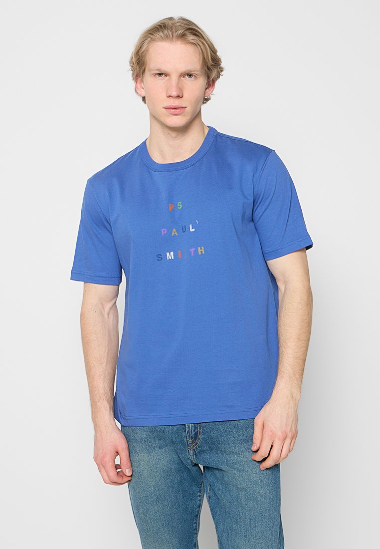 Junger Mann mit blonden Haaren, der ein blaues Paul Smith T-Shirt und blaue Jeans trägt und vor einem schlichten hellgrauen Hintergrund steht.