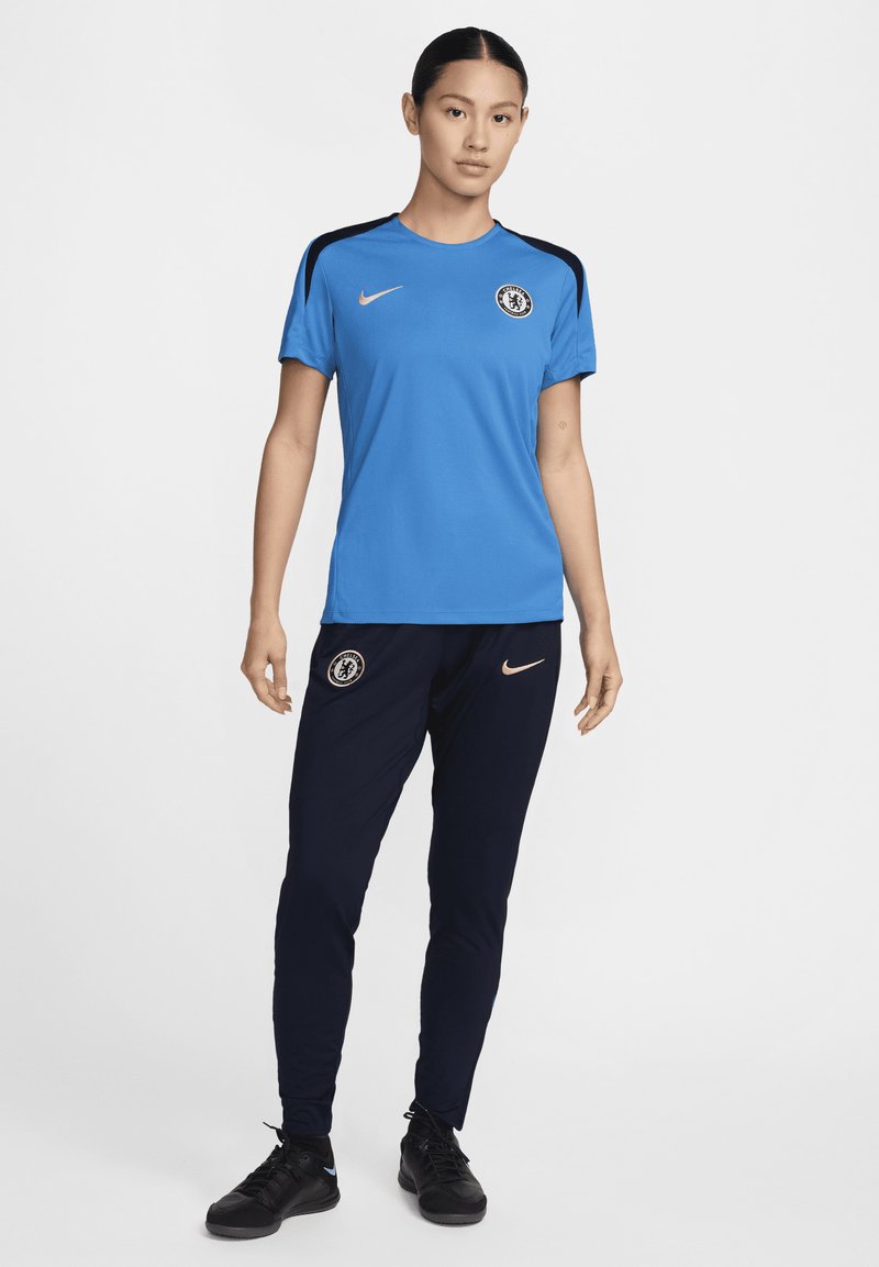 Blaues Sportshirt mit schwarzen Akzenten und Chelsea FC-Logo; kombiniert mit marineblauen Hosen mit Nike-Logo und schmalem Schnitt. Schwarze Sneakers.