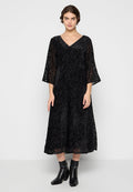 Schwarzes Spitzen-Midi-Kleid mit V-Ausschnitt und 3/4-Belledärmeln, das ein strukturiertes Blumenmuster aufweist. Getragen mit schwarzen Stiefeletten.