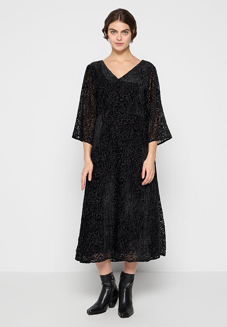 Schwarzes Spitzen-Midi-Kleid mit V-Ausschnitt und 3/4-Belledärmeln, das ein strukturiertes Blumenmuster aufweist. Getragen mit schwarzen Stiefeletten.