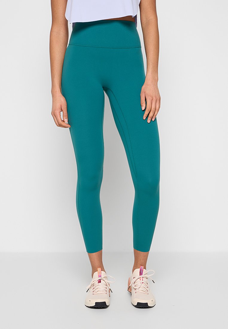 Frau trägt teal-farbene Leggings mit hohem Bund und hellrosa Sportschuhe, steht vor einem schlichten weißen Hintergrund.