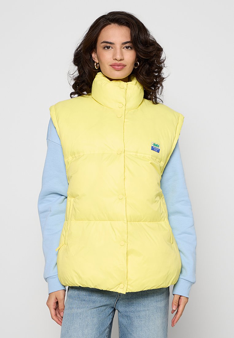 Gelbe Steppweste mit hohem Kragen, Druckknöpfen und Logo-Patch, getragen über einem hellblauen Langarmshirt. Glatte Textur, mittellange Form.