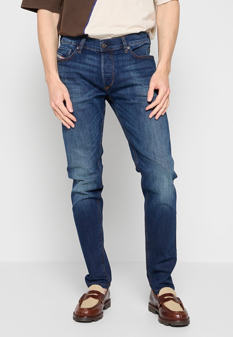 Mann trägt schmal geschnittene blaue Jeans mit leichten Abnutzungserscheinungen und Verfärbungen, kombiniert mit braunen und beigen Loafers, stehend vor einem schlichten Hintergrund.