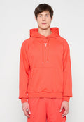 Mann trägt leuchtend orangefarbenen Kapuzenpullover und passende Hose, steht vor hellem, schlichtem Hintergrund und blickt nach vorne.