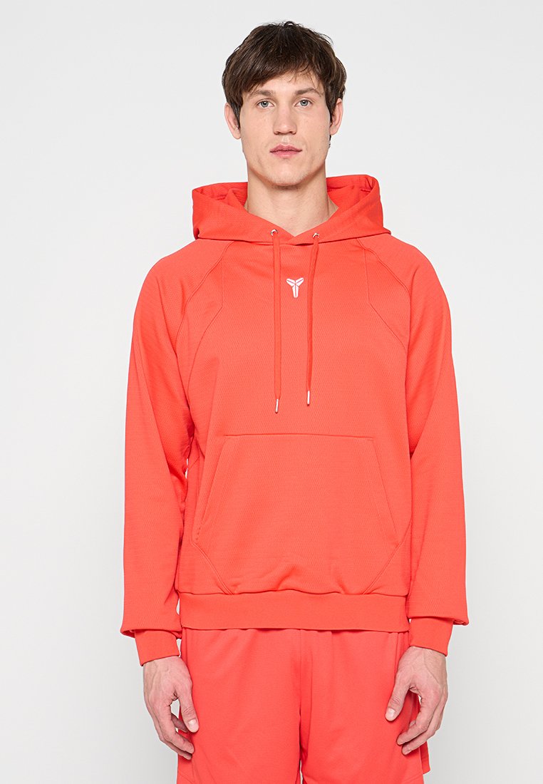 Mann trägt leuchtend orangefarbenen Kapuzenpullover und passende Hose, steht vor hellem, schlichtem Hintergrund und blickt nach vorne.